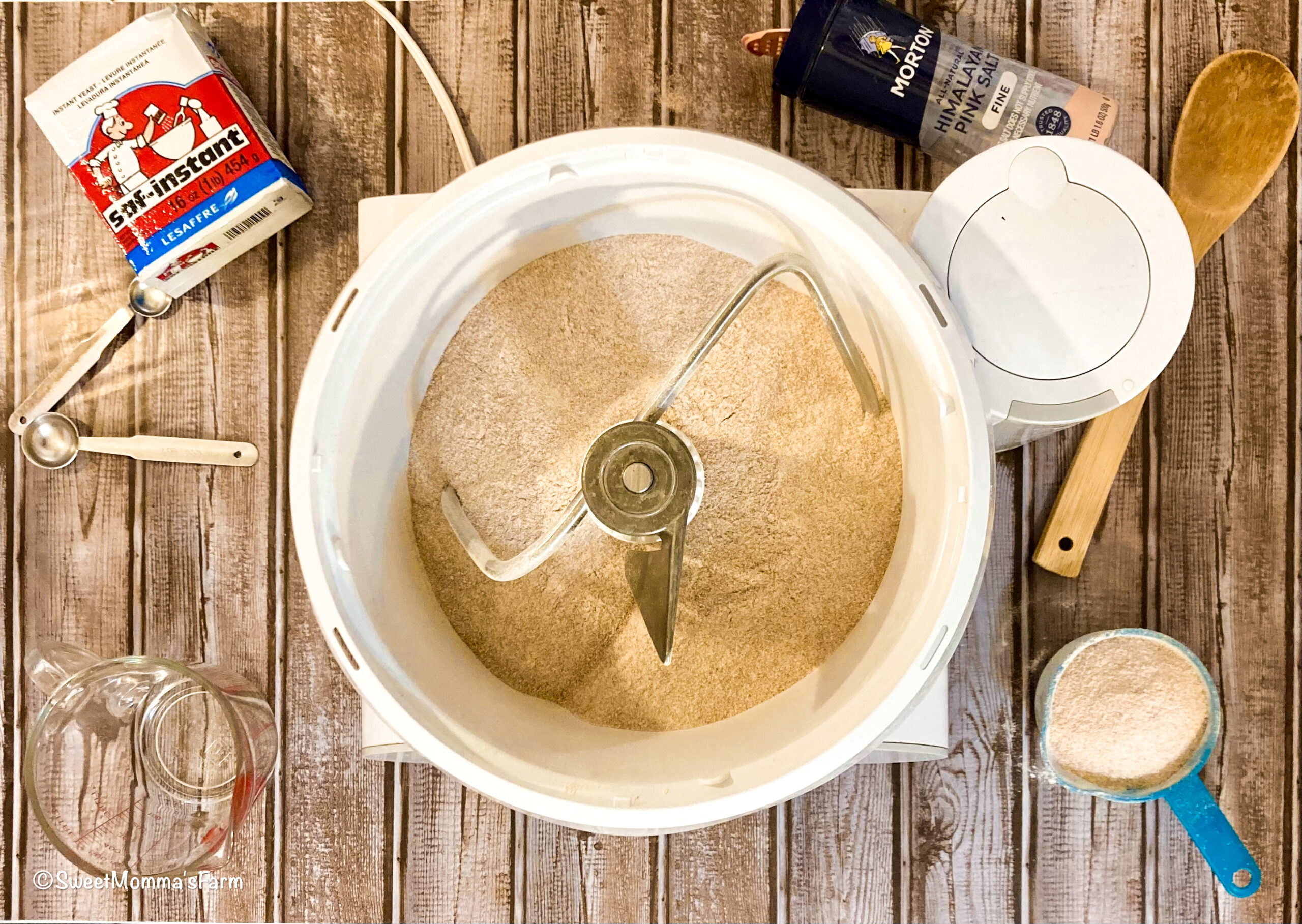 Mixer with bread hook. Salt, instant yeast, spoon, measuring cups and spoons lying around the mixture.