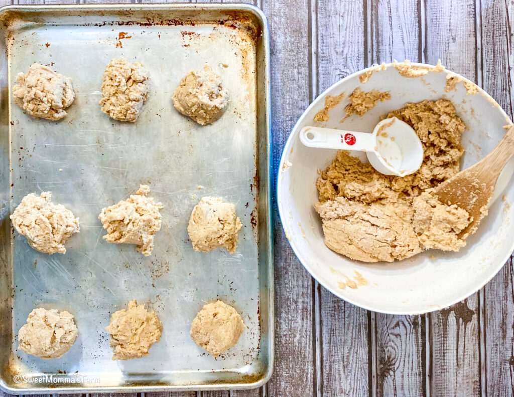 The biscuits dough is in a bowl with a spoon and a 1/4 measuring cup. The bowl is next to a cookie sheet with the biscuit dough dropped on to the pan